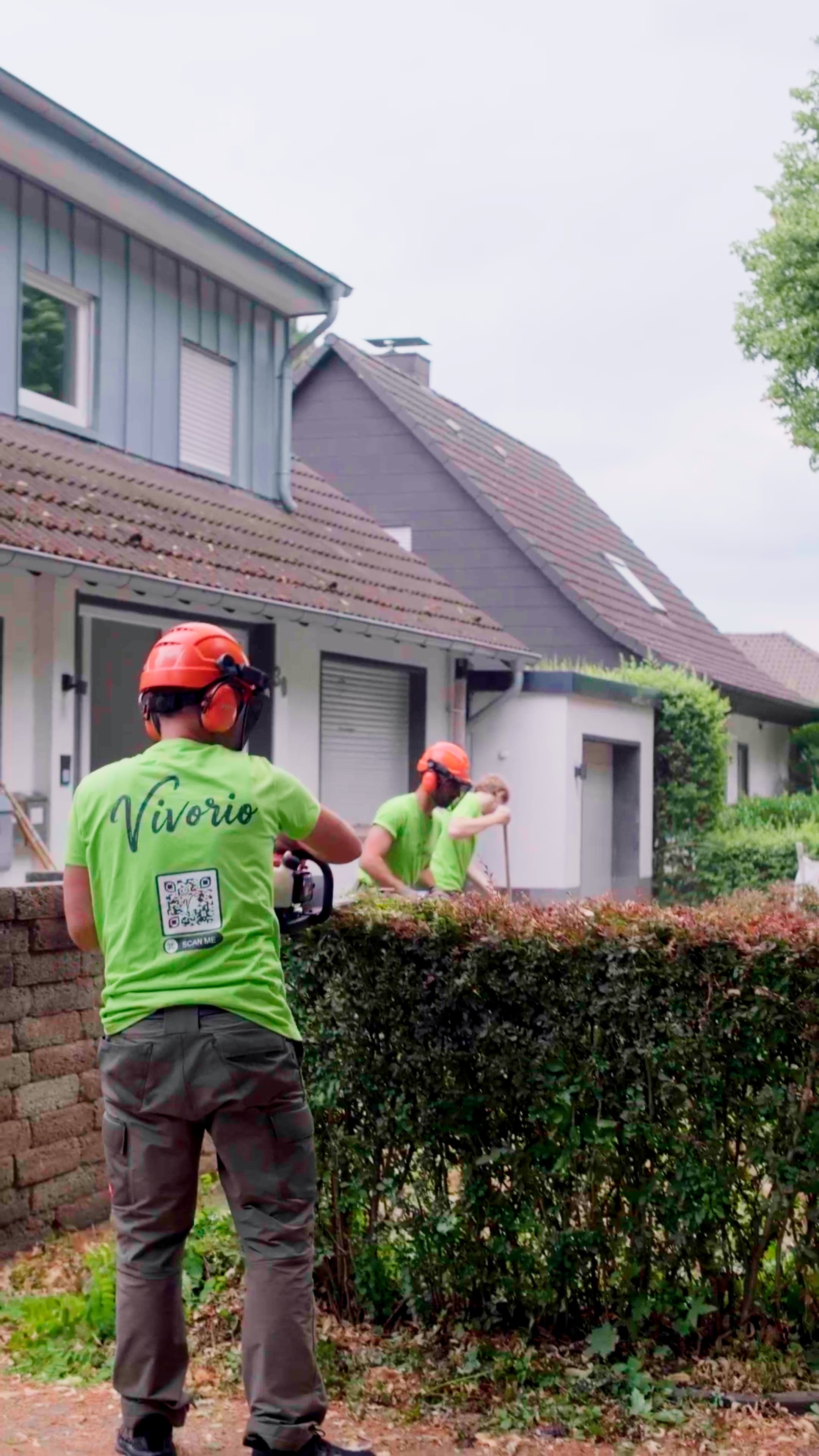 Zwei Mitarbeiter in hellgrünen Vivorio-Shirts schneiden eine Hecke vor Reihenhäusern.